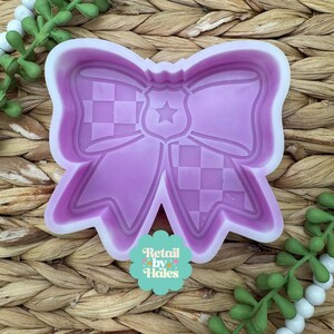 May include: A light purple silicone mold in the shape of a bow with a police badge design. The bow has a checkered pattern on the ends and a star in the center. The mold is surrounded by a white border and the text "Retail by Hales".