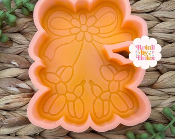 May include: An orange silicone mold with a floral and bow design. The mold features a large bow at the top, a flower in the center, and two sets of balloon-like shapes at the bottom. The text "Retail by Hales" is visible on the right side.