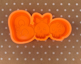 May include: Orange silicone cookie cutter shaped like the word "BOO" with a skull and bow in the "O".