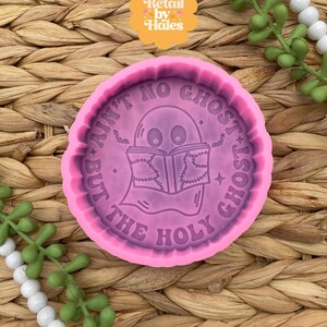 May include: A pink, circular silicone mold with the text "AIN'T NO GHOST BUT THE HOLY GHOST" surrounding a cartoon ghost reading a book. The mold is on a woven, brown surface with decorative greenery.