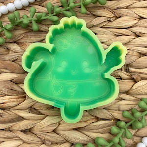 May include: A green silicone mold in the shape of a smiling cartoon character with arms and a floral crown. The mold has a light green outline and is surrounded by decorative green plant stems and white beads.