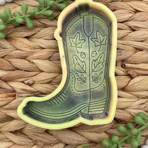 May include: A cowboy boot-shaped mold with a leaf design. The boot is outlined in black and filled with a muted green color. The mold has a yellow border and is set against a woven background.