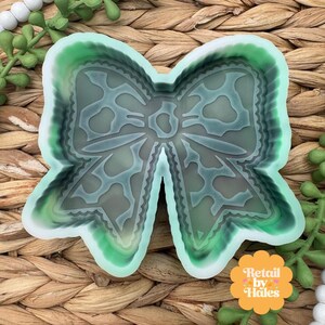 May include: A bow-shaped silicone mold with a translucent, light green and dark green marbled finish. The mold features a detailed, scalloped edge design and a central knot area, providing a clear, hollow cavity for crafting purposes.