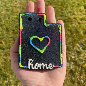 Peut inclure: Ornement en paillettes noires en forme de l'&eacute;tat de l'Utah avec un contour vert n&eacute;on, rose et bleu. L'ornement a un c&oelig;ur blanc au centre et le mot "home" &eacute;crit en blanc sous le c&oelig;ur.