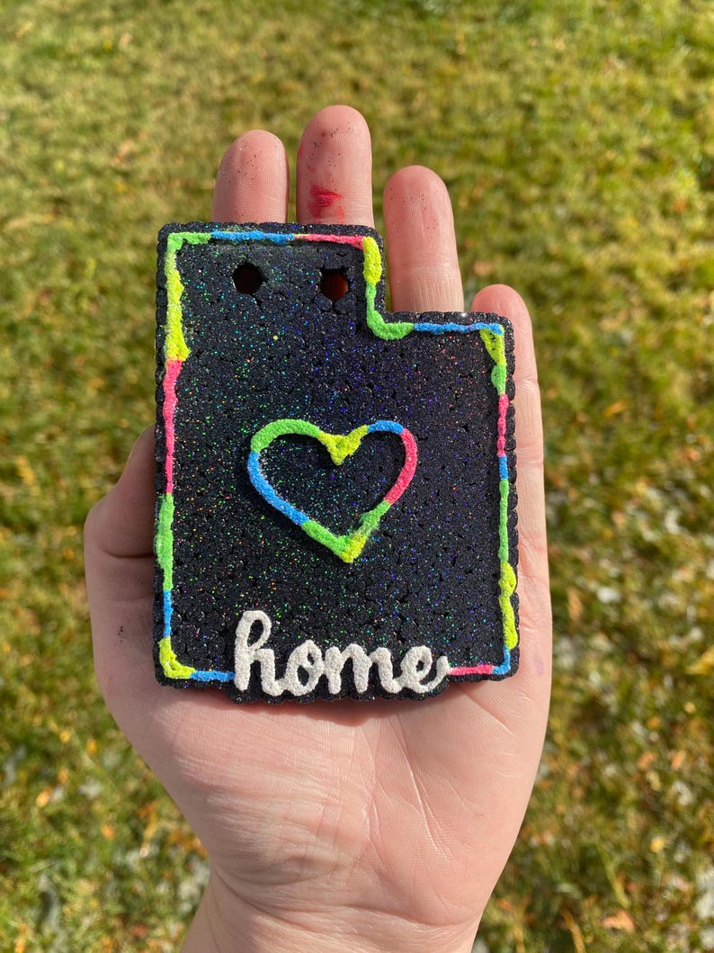 Peut inclure: Ornement en paillettes noires en forme de l'&eacute;tat de l'Utah avec un contour vert n&eacute;on, rose et bleu. L'ornement a un c&oelig;ur blanc au centre et le mot "home" &eacute;crit en blanc sous le c&oelig;ur.
