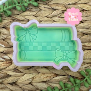 May include: A light pink and green silicone mold in the shape of stacked books with bows. The books are green with a checkered pattern in the middle. The mold has the text "Retail by Hales" in a pink flower shape.