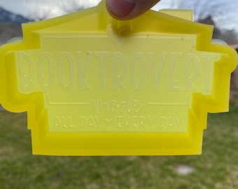 May include: Yellow plastic cookie cutter shaped like a house with the text "Booktrovert mode all day every day" and a heart symbol.