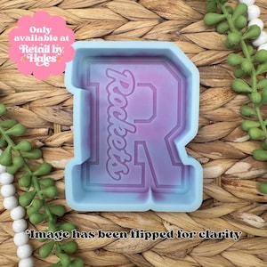 May include: A light blue silicone mold in the shape of the letter R with a purple interior. The mold features the word Rockets embossed in a script font within the letter shape. The mold is designed for casting and has a smooth finish.