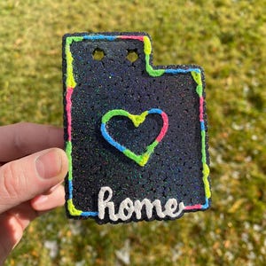 Peut inclure: Une pancarte de d&eacute;coration d'int&eacute;rieur noire et scintillante avec un contour vert n&eacute;on, rose et bleu. La pancarte a la forme de l'&eacute;tat de l'Utah et comporte un c&oelig;ur au centre avec le mot "home" &eacute;crit en dessous.