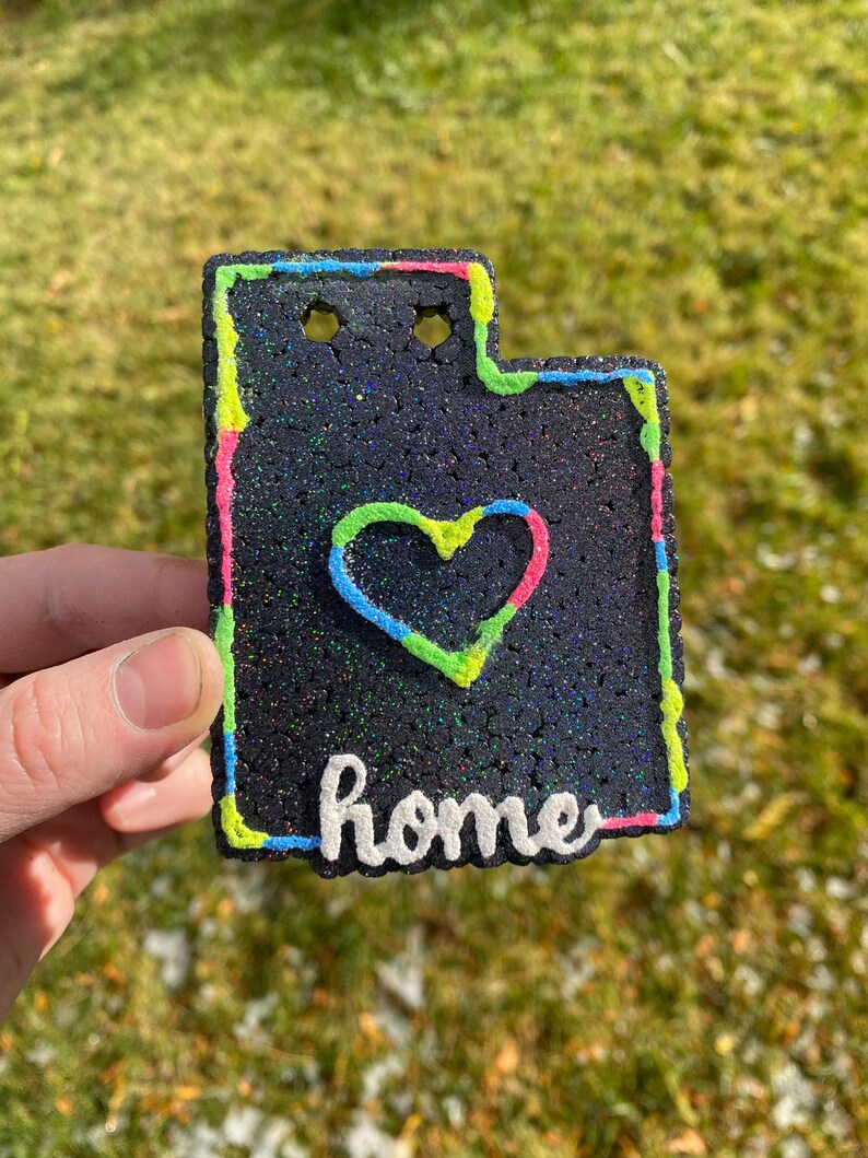 Peut inclure: Une pancarte de d&eacute;coration d'int&eacute;rieur noire et scintillante avec un contour vert n&eacute;on, rose et bleu. La pancarte a la forme de l'&eacute;tat de l'Utah et comporte un c&oelig;ur au centre avec le mot "home" &eacute;crit en dessous.