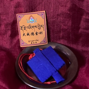 May include: An orange card with ornate borders and text in a foreign language sits atop a small, dark bowl. A blue fabric item with a red cord is inside the bowl. The background is a deep red fabric.