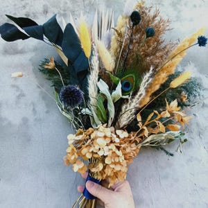 May include: A dried flower bouquet featuring a peacock feather, blue thistle, and various dried grasses and leaves. The bouquet includes yellow, brown, and green elements, tied with a blue ribbon. The arrangement is held in a person's hand.