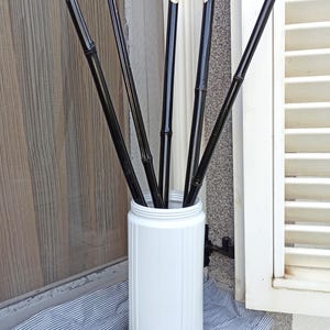 Op de afbeelding: Een witte cilindervormige vaas met een bruine basis bevat verschillende zwarte bamboestengels. Een groene limoen ligt op een gestreepte blauw-witte stof voor de vaas, bij een raam.