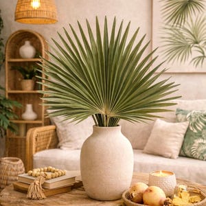 Könnte beinhalten: Eine große, hellfarbene Vase enthält eine fächerförmige Palmenblattanordnung. Die Vase steht auf einem Holztisch mit Büchern, Perlen und einem kleinen Korb mit Obst. Der Hintergrund umfasst ein Sofa, Kissen und eine Wand mit einem gerahmten botanischen Druck.