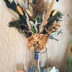 May include: A dried flower bouquet in a clear glass vase. The bouquet includes peacock feathers, dried hydrangeas, and various grasses. The vase is tied with a blue ribbon and sits on a white surface with a candle and a decorative house.
