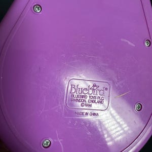 Puede incluir: Un componente de juguete de pl&aacute;stico morado con forma redondeada, asegurado por cuatro tornillos. La etiqueta dice "Bluebird Toys PLC, Swindon, England &copy;1994 Made in China."