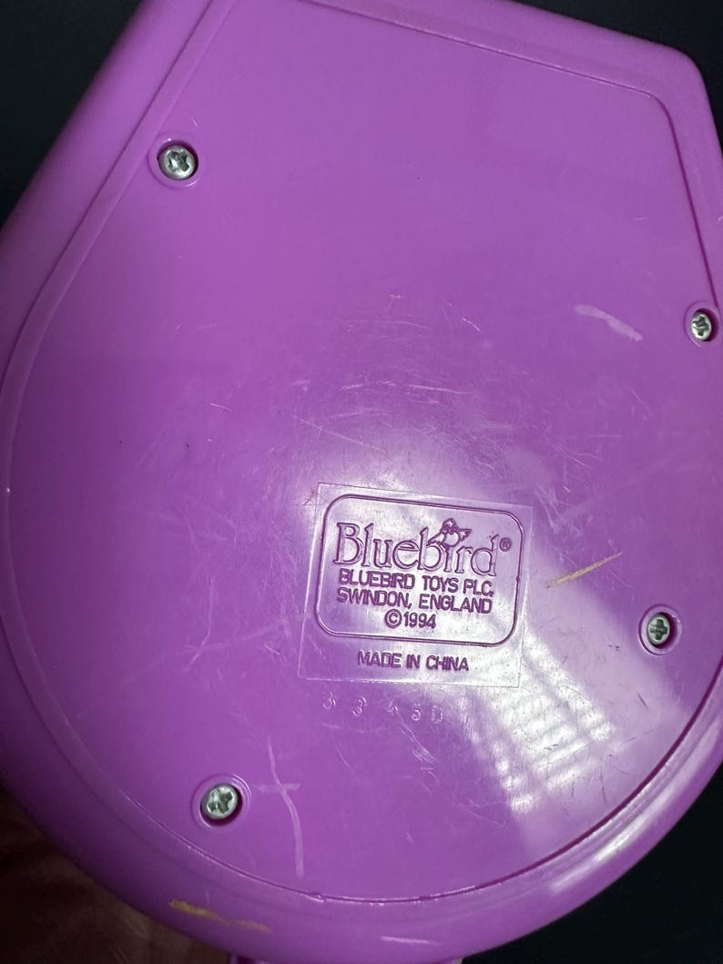 Puede incluir: Un componente de juguete de pl&aacute;stico morado con forma redondeada, asegurado por cuatro tornillos. La etiqueta dice "Bluebird Toys PLC, Swindon, England &copy;1994 Made in China."