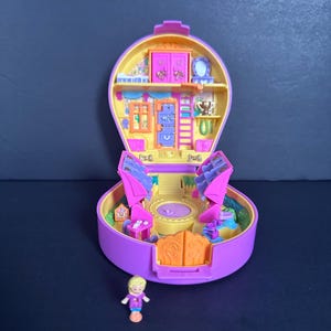 Puede incluir: Un juego de Polly Pocket vintage morado y amarillo, abierto para revelar un interior en miniatura con muebles y una peque&ntilde;a mu&ntilde;eca. El juego presenta un dise&ntilde;o redondo con una abertura con bisagras, mostrando un mundo en miniatura detallado.