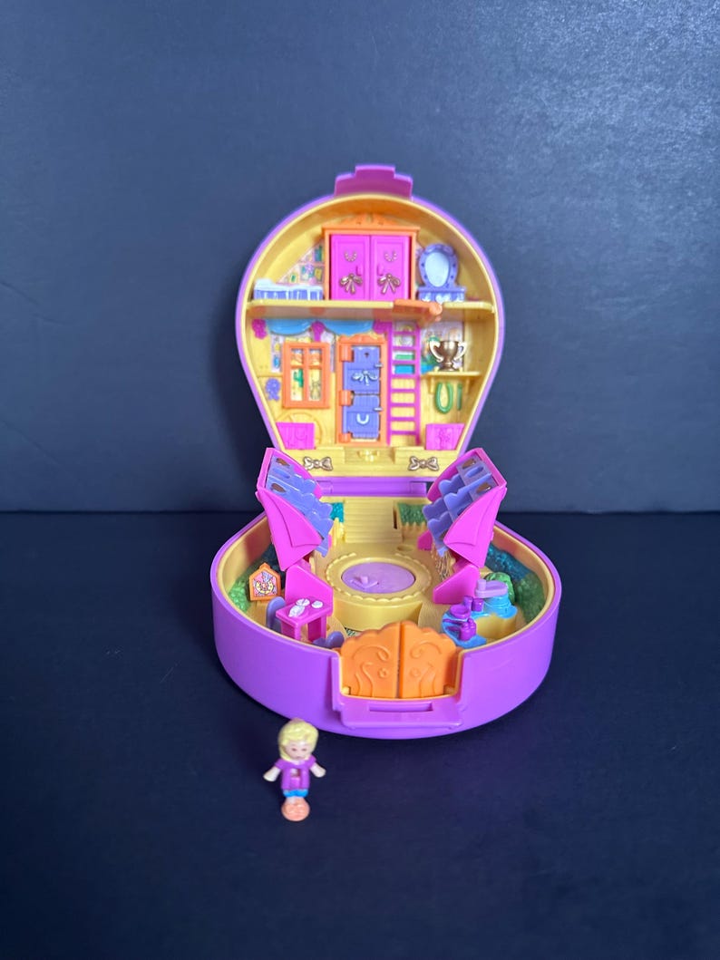 Puede incluir: Un juego de Polly Pocket vintage morado y amarillo, abierto para revelar un interior en miniatura con muebles y una peque&ntilde;a mu&ntilde;eca. El juego presenta un dise&ntilde;o redondo con una abertura con bisagras, mostrando un mundo en miniatura detallado.