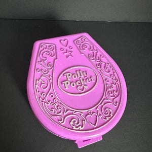 Puede incluir: Un compacto Polly Pocket vintage en un vibrante color rosa. El compacto tiene forma de herradura con detalles ornamentados en color plata y las palabras "Polly Pocket" en el centro. Tambi&eacute;n hay corazones y estrellas decorativas.