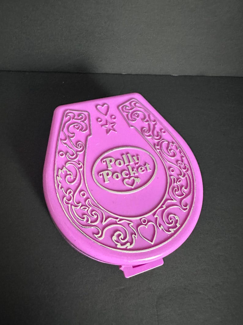 Puede incluir: Un compacto Polly Pocket vintage en un vibrante color rosa. El compacto tiene forma de herradura con detalles ornamentados en color plata y las palabras "Polly Pocket" en el centro. Tambi&eacute;n hay corazones y estrellas decorativas.