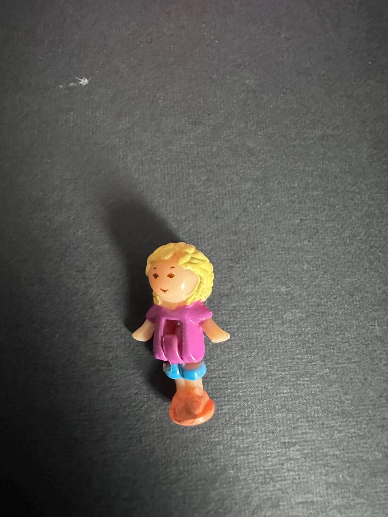 Puede incluir: Peque&ntilde;a mu&ntilde;eca de pl&aacute;stico con pelo rubio, top rosa, pantalones cortos azules y zapatos naranjas. La mu&ntilde;eca tiene una cara sonriente y los brazos extendidos. Es una figura en miniatura, probablemente para jugar o coleccionar.