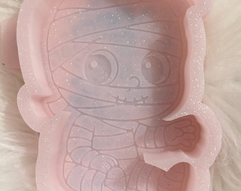 May include: A light pink, glittery silicone mold in the shape of a cartoon mummy. The mummy has large eyes, a stitched mouth, and is wrapped in bandages. The mold is designed for crafting and creating resin or other decorative items.