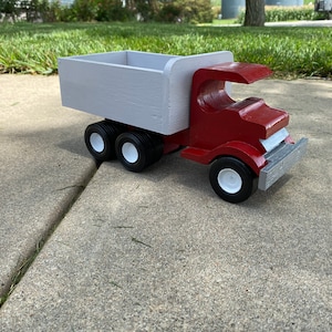 May include: A red toy dump truck with a white wooden bed. The truck has black wheels with white rims and a gray bumper.
