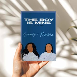 Pode incluir: Uma impressão retangular azul com o texto "THE BOY IS MINE TOUR" e os nomes "Brandy + Monica" em escrita branca. A impressão apresenta ilustrações estilizadas de duas mulheres. A impressão é segurada em uma mão.