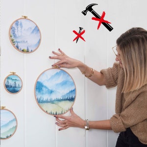 May include: A woman is hanging a set of four round wood framed watercolor paintings on a white wall. The paintings feature a landscape scene with mountains, trees, and fog. The paintings are different sizes.
