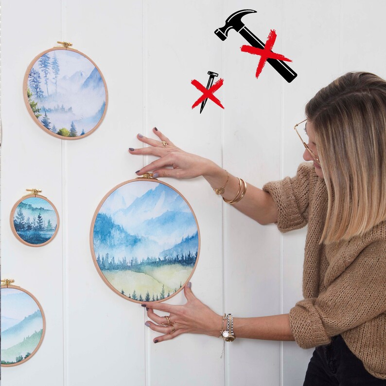 May include: A woman is hanging a set of four round wood framed watercolor paintings on a white wall. The paintings feature a landscape scene with mountains, trees, and fog. The paintings are different sizes.