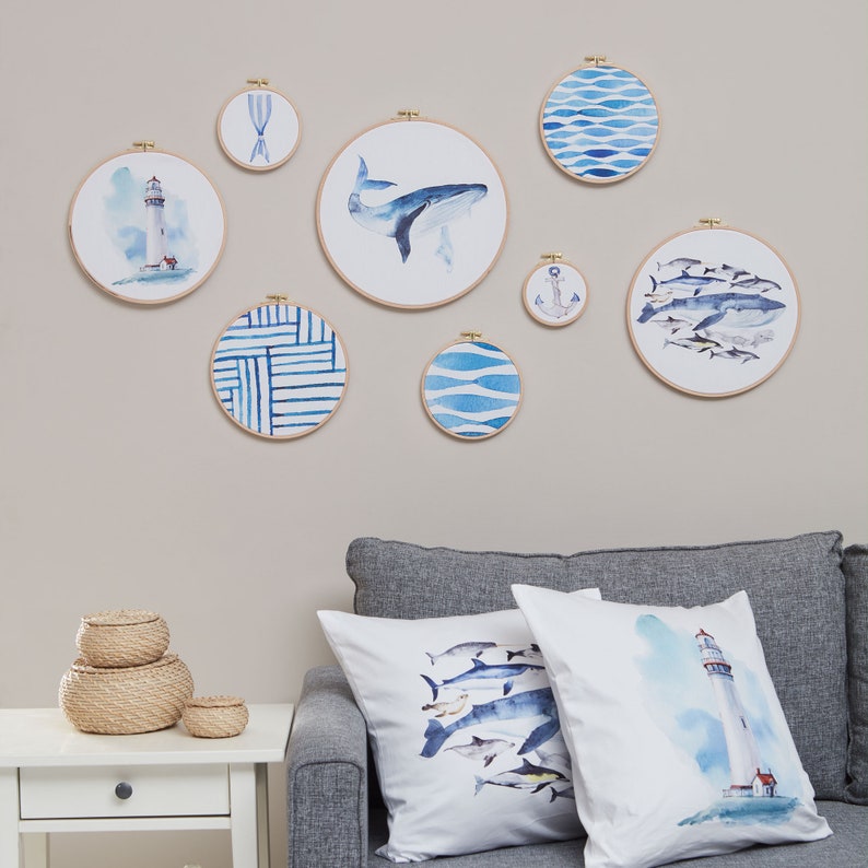 May include: A collection of eight blue and white watercolor embroidery hoops with nautical themes, including a lighthouse, whale, anchor, and fish. The hoops are displayed on a light beige wall above a gray couch with two white throw pillows featuring watercolor designs of a lighthouse and a school of fish.