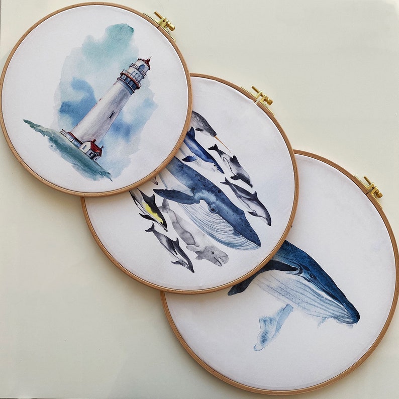 May include: Three embroidery hoops with watercolor paintings. The first hoop features a lighthouse on a blue and green background. The second hoop features a whale and other sea creatures on a white background. The third hoop features a whale on a white background.