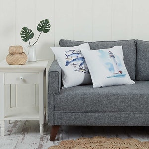 May include: Two decorative throw pillows with watercolor designs. One pillow features a lighthouse with a blue sky and water. The other pillow features a school of fish with a white background.