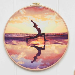 Puede incluir: Un aro de bordado redondo con una imagen impresa de una persona haciendo yoga en una playa al atardecer. La persona est&aacute; silueteada contra el cielo naranja y rosa, y su reflejo es visible en el agua.