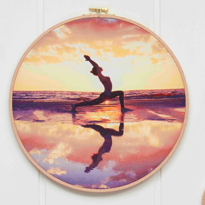Puede incluir: Un aro de bordado redondo con una imagen impresa de una persona haciendo yoga en una playa al atardecer. La persona est&aacute; silueteada contra el cielo naranja y rosa, y su reflejo es visible en el agua.