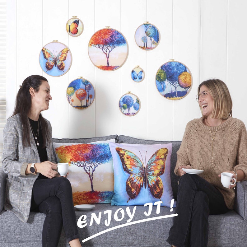 Puede incluir: Una sala de estar con un sof&aacute; y dos cojines con dise&ntilde;os de mariposas. Los cojines est&aacute;n frente a una pared blanca con varios aros de bordado enmarcados con dise&ntilde;os de mariposas y &aacute;rboles. El texto "enjoy it!" est&aacute; escrito en blanco en el sof&aacute;.