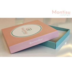 Puede incluir: Una caja rosa con un logotipo verde y blanco que dice "Montisu uniquely designed" y un nombre de usuario de Instagram blanco que dice "@montisuofficial". La caja est&aacute; abierta y el interior es de color azul claro.