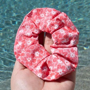 Può includere: Un elastico per capelli color corallo con un motivo floreale bianco. Il tessuto è arricciato ed elastico, creando una forma circolare. L'elastico per capelli è tenuto in una mano, con uno sfondo d'acqua sfocato.