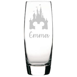 May include: A clear glass champagne flute with an etched design of a castle and Mickey Mouse ears. The name "Emma" is etched below the design.