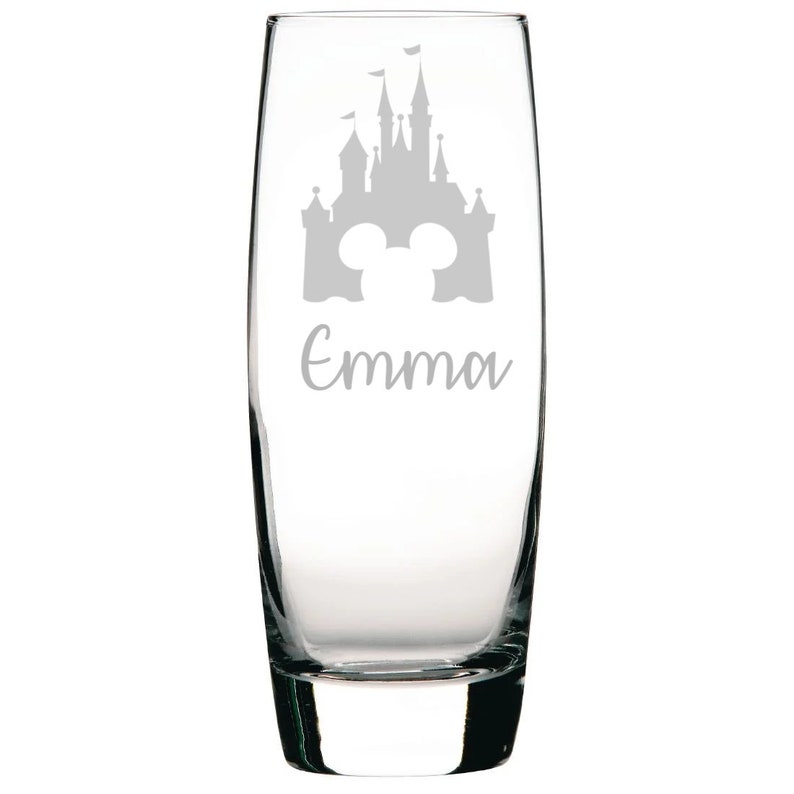 May include: A clear glass champagne flute with an etched design of a castle and Mickey Mouse ears. The name "Emma" is etched below the design.