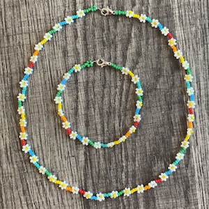 Könnte beinhalten: Zwei Perlenketten mit weißen Gänseblümchen und bunten Perlen. Die größere Kette ist rot, orange, gelb, grün, blau und lila. Die kleinere Kette ist grün, blau und gelb.