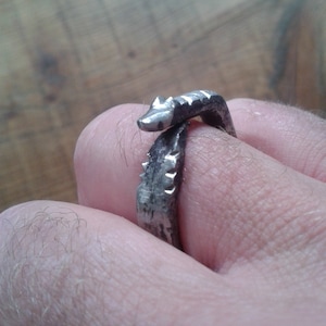May include: A silver metal ring shaped like a snake with a textured surface.