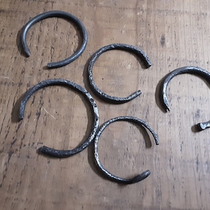 Peut inclure: Cinq bracelets en métal ouverts, fabriqués à la main, avec une finition sombre et texturée. Les bracelets sont disposés sur une surface en bois, mettant en valeur leur design simple mais rustique. Chaque bracelet a une forme et une texture uniques.