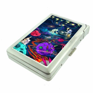 May include: A silver cigarette case with a colorful space-themed design featuring astronauts, planets, and stars. The case has a hinged lid and a push button to open.
