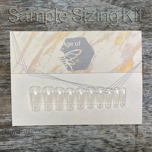 Może przedstawiać: Biała karta z nadrukiem "Sample Sizing Kit" i "Age of e. Age of Be.". Na karcie znajduje się rząd dziesięciu przezroczystych plastikowych końcówek do paznokci ponumerowanych od 0 do 9.
