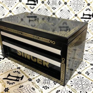 May include: Black Singer sewing machine cabinet with gold accents. The cabinet has two silver-colored drawers and the Singer logo in gold. The cabinet is rectangular and sits on a patterned fabric.
