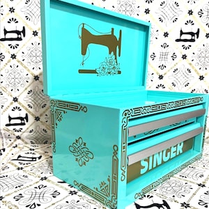 May include: A turquoise sewing box with a gold Singer logo and decorative accents. The box features a gold silhouette of a sewing machine and floral details on the lid. The front of the box has the word "SINGER" in gold.