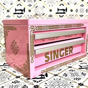 May include: A pink Singer sewing box with two drawers and gold accents. The word "SINGER" is prominently displayed in gold lettering on the front. The box has decorative gold trim and a patterned design.