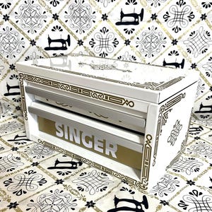 May include: A white Singer sewing machine storage box with gold and silver accents. The box has a gold panel with the Singer logo in silver. The box is decorated with gold filigree and sits on a patterned fabric.